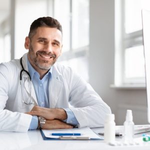 portrait-of-happy-male-doctor-in-white-medical-uniform-sitting-at-desk-in-hospital-and-smiling-at-e1736272190315.jpg